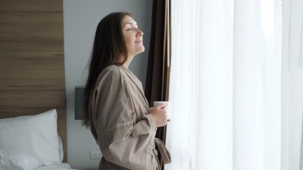 Κορίτσι με μπουρνούζι πίνει καφέ κοντά στο παράθυρο στο δωμάτιο του ξενοδοχείου — Αρχείο Βίντεο