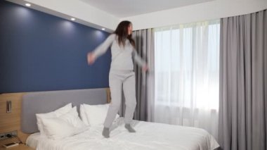 Neşeli kız otel odasındaki pencereye karşı büyük bir yatakta zıplıyor.