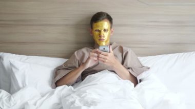 Altın maskeli adam yumuşak yatakta uzanan akıllı telefonu kullanıyor.