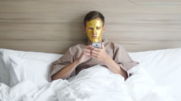 Uomo con maschera viso dorata utilizza smartphone sdraiato nel letto morbido — Video Stock