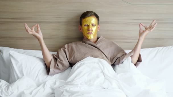 Man in coat with golden face mask meditates in bed in hotel — Stock Video