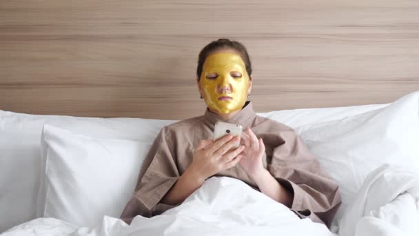 Chica con máscara dorada utiliza teléfono inteligente en la cama suave — Vídeos de Stock