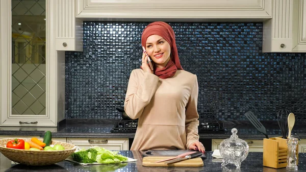 Mulher no hijab fala no smartphone em pé na mesa da cozinha — Fotografia de Stock