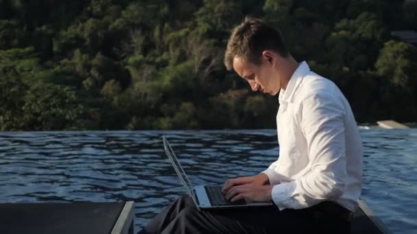 Uomo d'affari lavora sul computer portatile contro foresta e piscina dell'hotel — Video Stock