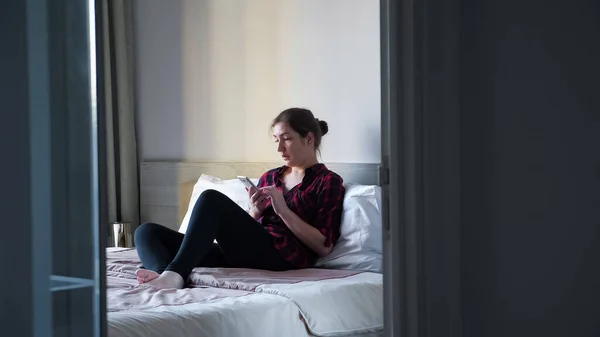 Mujer joven se sienta en una cama grande con almohadas blancas y tipos en el teléfono inteligente en el dormitorio —  Fotos de Stock