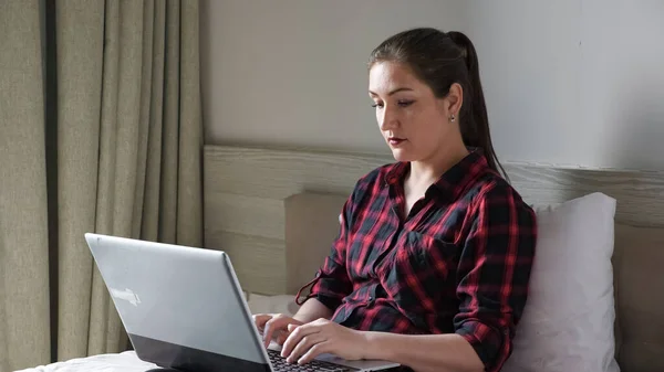 예쁜 여자가 갈색 침대에 앉아 현대 회색 노트북의 타입 — 스톡 사진