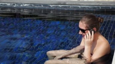 Güneş gözlüklü ve mayo giymiş genç bir kadın akıllı telefondan konuşuyor.