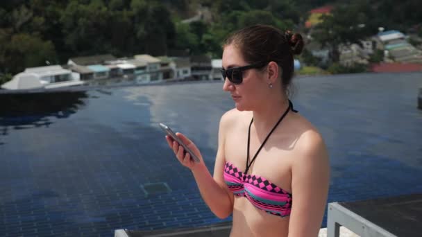 Mladá žena typy na černém smartphone s úsměvem v blízkosti bazénu — Stock video