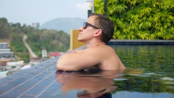Красивий чоловік в сонцезахисних окулярах спирається на готельний край басейну — стокове відео
