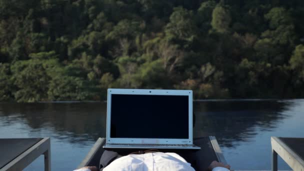 Notebook s tmavou obrazovkou na mužských nohách proti rostlinám a bazénu — Stock video