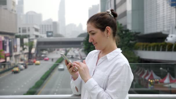 Брюнетка в белых типах на смартфоне на мосту через улицу — стоковое видео