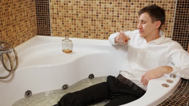 Hombre borracho en pantalones y camisa está acostado en un baño con agua, beber alcohol — Vídeos de Stock