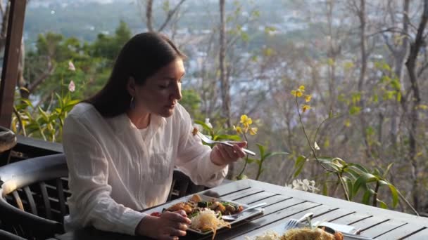 Woman in white shirt eats noodles with vegetables at an outdoor cafe — Stock Video