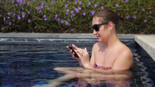 Menina em preto óculos de sol tipos no smartphone sentado na piscina — Vídeo de Stock