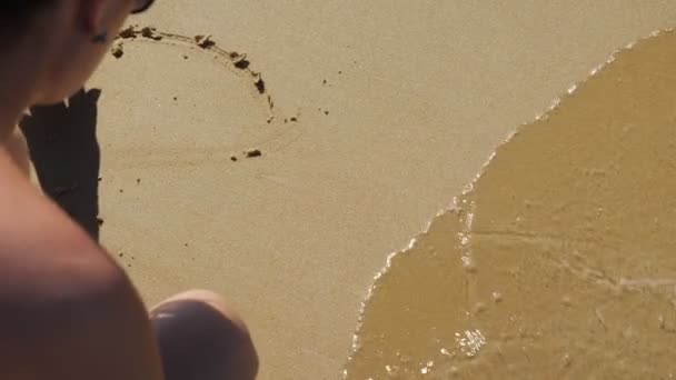 Jonge vrouw tekent een hart op het zand. Zeegolf nadert — Stockvideo