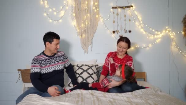 Szczęśliwi rodzice na łóżku przeciwko girlandzie żarówki na ścianie — Wideo stockowe