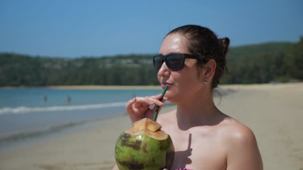 Güneş gözlüklü genç kadın sahilde hindistan cevizi içiyor. — Stok video