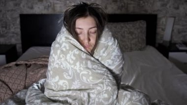 Genç bir kadın battaniyeye sarılmış yatağın kenarında oturuyor.