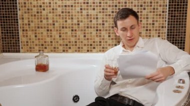 Pantolon ve gömlekli sarhoş adam banyoda uzanıyor ve belgeleri inceliyor.