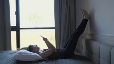 Gülümseyen kız yatakta otelde pencereye karşı internette sörf yapıyor.