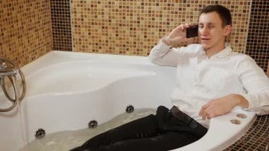 Pantolon ve gömlek giyen bir adam su banyosunda yatıyor, telefonda konuşuyor.