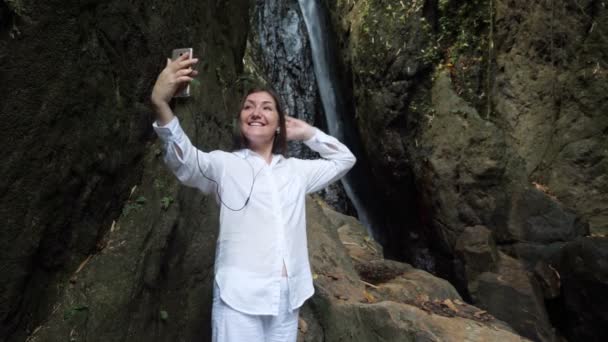Vrouw in hoofdtelefoon voert videogesprek tegen waterval — Stockvideo