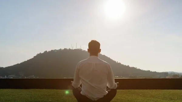 Man meditates on grass on sunny day against big green hill — 스톡 사진