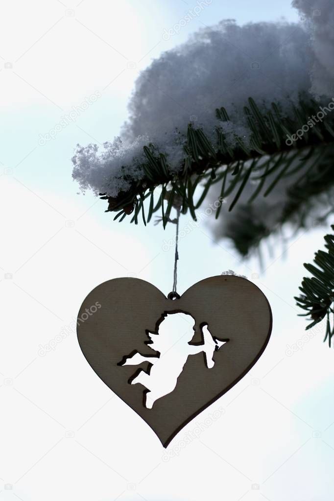 Cupid silhouette on heart shape wooden decoration, hanging on pine branch covered by snow, isolated on sky background.