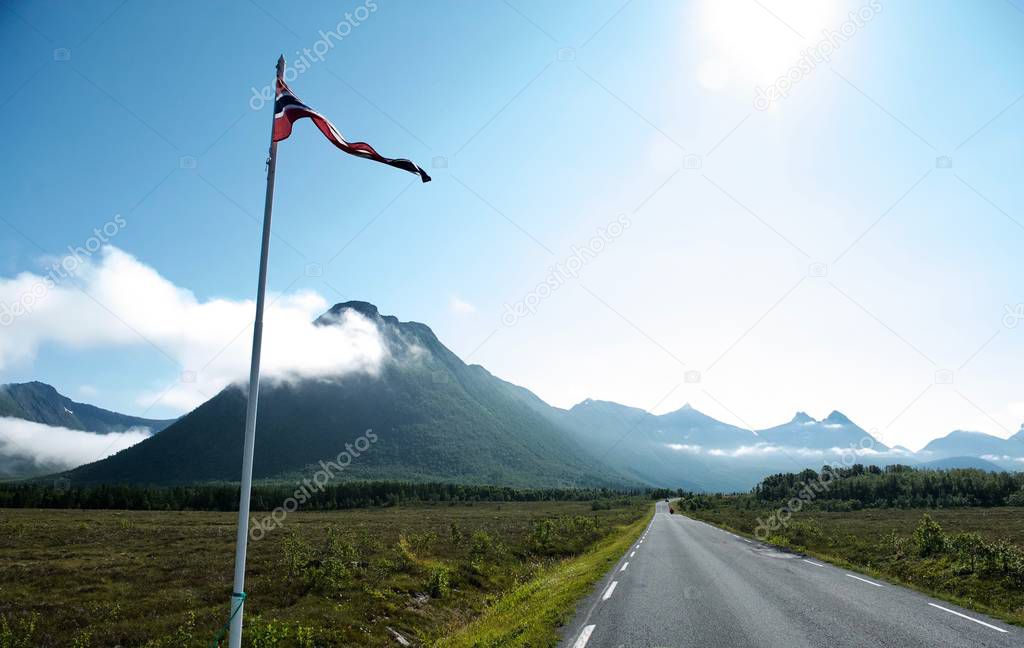 Straight road through the green meadows to the mountains. Sun shining in the sky up on road and mountains . Norway flag on the left side of the road on the foregroung of picture.