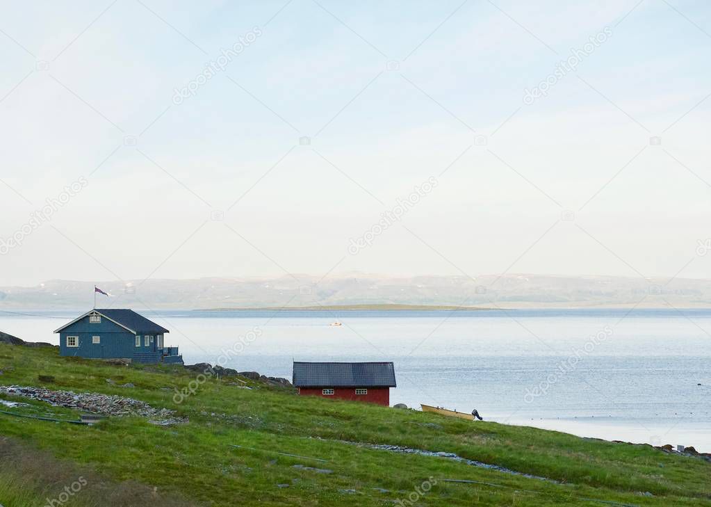 Traditional red and grey colored fishing houses with Norway flag and boat on the fjord coastline. Polar day. Nighttime.