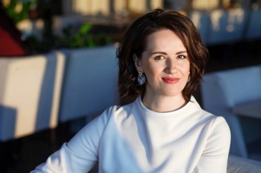 Bir kafede açık renk elbise giymiş mutlu güzel bir kadının portresi. Başarılı, olgun bir kadın gülümseyen ve kendinden emin bir şekilde kameraya bakıyor.. 