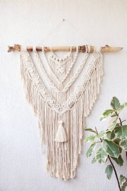 Handmade macrame  100% cotton wall decoration with wooden stick hanging on a white wall.  Modern decoration in the interior. Macrame braiding and cotton threads.  Female hobby.   clipart