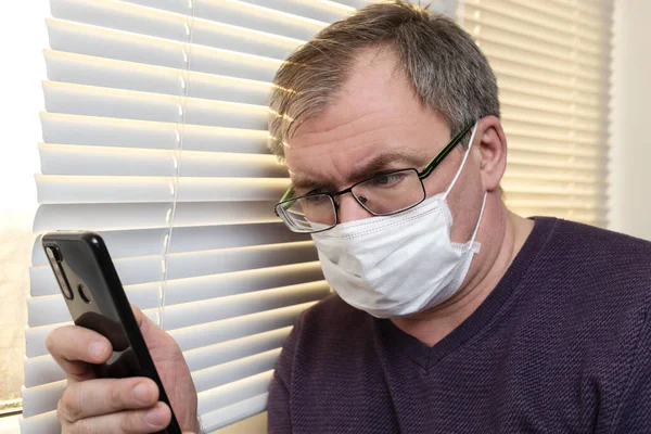 A man in a medical mask looks at a smartphone against the background of blinds. Self-isolation mode. Stay at home