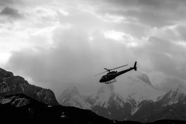 An atmospheric picture shows the silhouette of the helicopter climbing in front of the high snowy mountains.