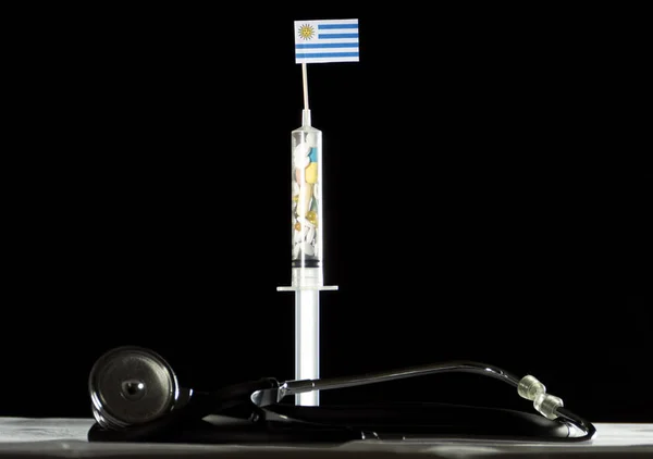 Estetoscopio y jeringa llenos de drogas inyectando la bandera uruguaya sobre fondo negro — Foto de Stock