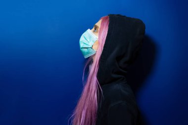 Studio profile portrait of young girl with pink hair and blue eyes, wearing medical flu mask and hooded sweater, looking up, on background of phantom blue color.