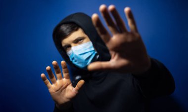 Kapüşonlu kazak giyen, tıbbi grip maskesi takan genç bir adamın stüdyo portresi. Dur işaretini elleriyle gösteriyor. Koyu mavi arkaplanda izole.