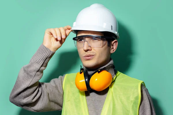 stock image Studio portrait of young builder man, engineer, touching his safety helmet for construction, wearing glasses and headphones on background of aqua menthe color.