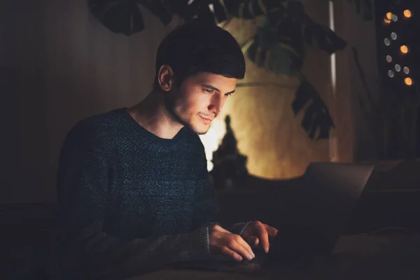Night portrait of young cheerful guy working on laptop in dark room with garlands on table at home.