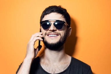 Akıllı telefondan konuşan, turuncu arka planda güneş gözlüğü takan genç bir adamın stüdyo portresi..