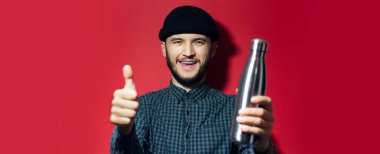 Mutlu genç bir adamın stüdyo portresi, kırmızı arka planda tekrar kullanılabilir çelik termo su şişesini gösteriyor ve tutuyor..