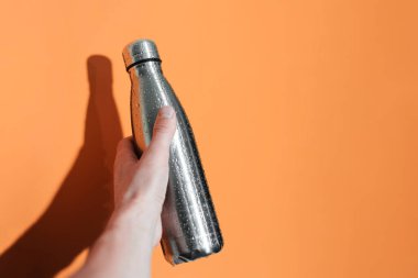 Erkek elinin, herhangi bir sıvı için tekrar kullanılabilir çelik bir termo eko şişeyi tutarken, su püskürtülmüş, turuncu arka planda izole edilmiş..