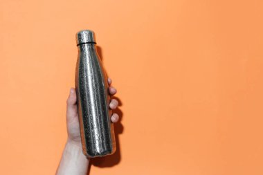 Erkek elinin, herhangi bir sıvı için tekrar kullanılabilir çelik bir termo eko şişeyi tutarken, su püskürtülmüş, turuncu arka planda izole edilmiş..