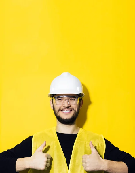 若い笑顔の男の肖像 ビルダーエンジニア 親指を表示 建設のための安全ヘルメットを身に着けています 眼鏡と黄色のジャケット 黄色の背景に — ストック写真