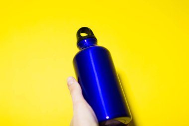 Elinde alüminyum termo eko su şişesi var. Klasik mavi renk, sarı arka planda izole edilmiş..