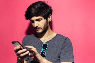 Genç hippi portresi, gömlek üzerine güneş gözlüğü asmak, akıllı telefon ve kablosuz kulaklık kullanmak, pembe mercan arka planda izole edilmiş..