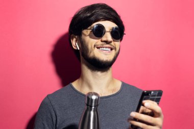 Güneş gözlüklü, kablosuz kulaklık ve akıllı telefon kullanan, mercan pembesi arka planında çelik termodinamik su şişesi tutan genç bir adamın portresi..