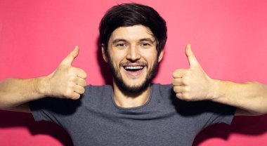 Genç mutlu adamın portresi mercan pembe arka planda izole edilmiş baş parmağını gösteriyor..