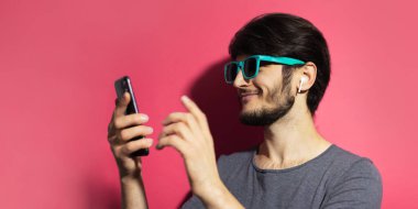 Akıllı telefon ve kablosuz kulaklık kullanan, pembe mercan arka planda Cyan güneş gözlüğü takan genç bir adamın portresi..