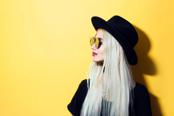 Portrait of young hipster blonde girl, looking in other side, wearing black hat, sunglasses and shirt, isolated on yellow background.